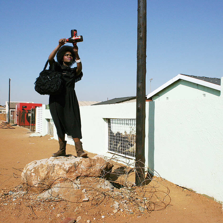 a black young woman in a black oldfashioned dress is standing on a big stone on the side of a yellow dusty street, she is holding up two bottles of black lalbel beer in the form of a cross into the blue sky, the stone is secured by barbwire