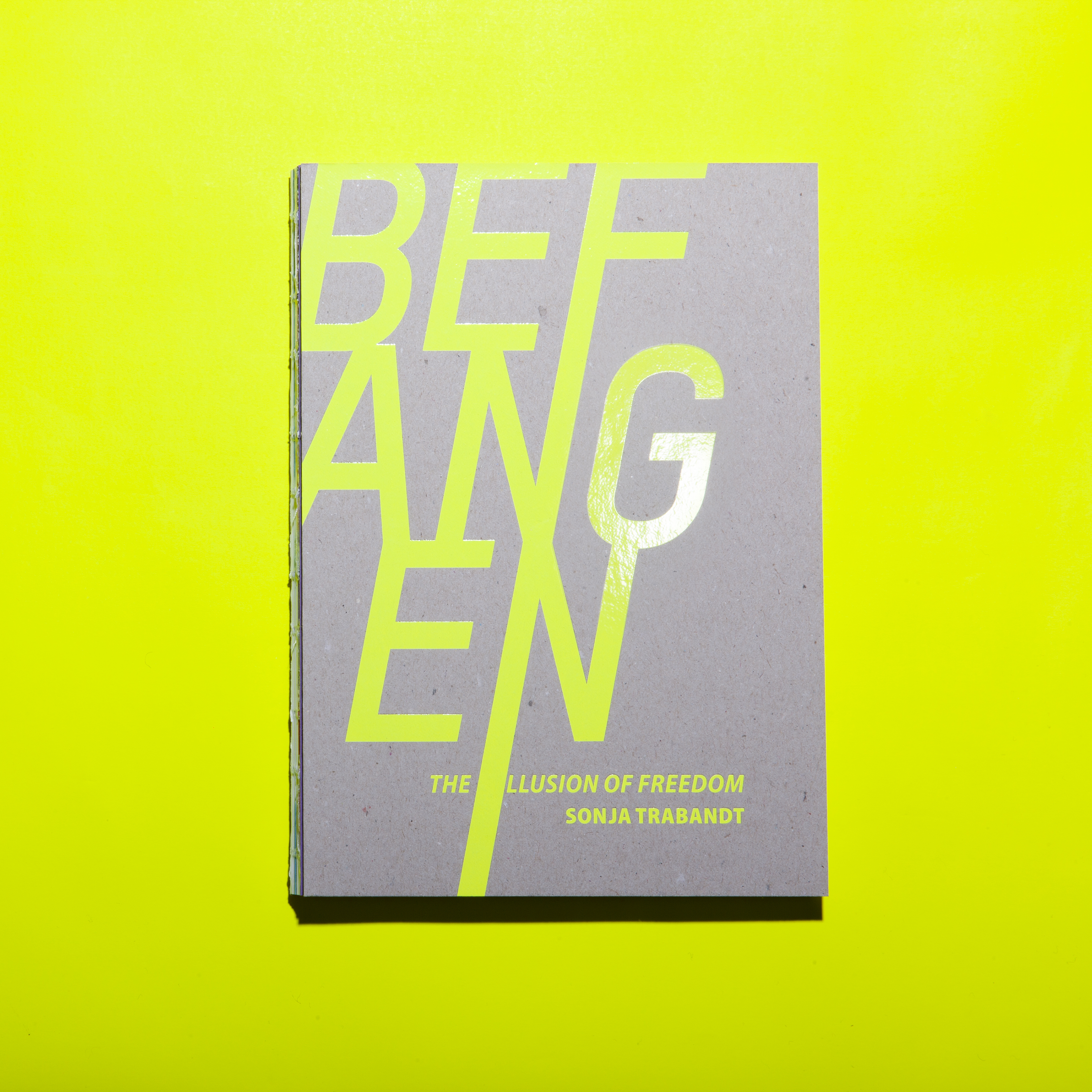 cover of the photobook befangen-the illusion of freedom, it is neon yellow print of the title and author Sonja Trabandt on grey cardboard with an open binding